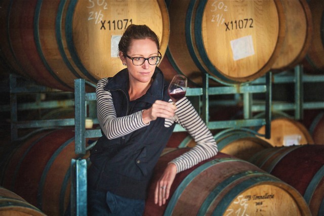A woman stands holding a wine glass in her right hand while here left arm leans on a barrel.