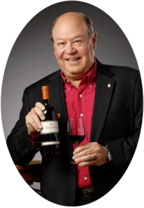 Bob Monica in a black blazer and red collared shirt holding a bottle of wine in his right hand and a glass of wine in his left.