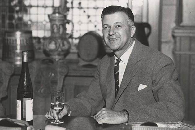 John Daniel, Jr. sits at a table with a glass of wine in his right hand.
